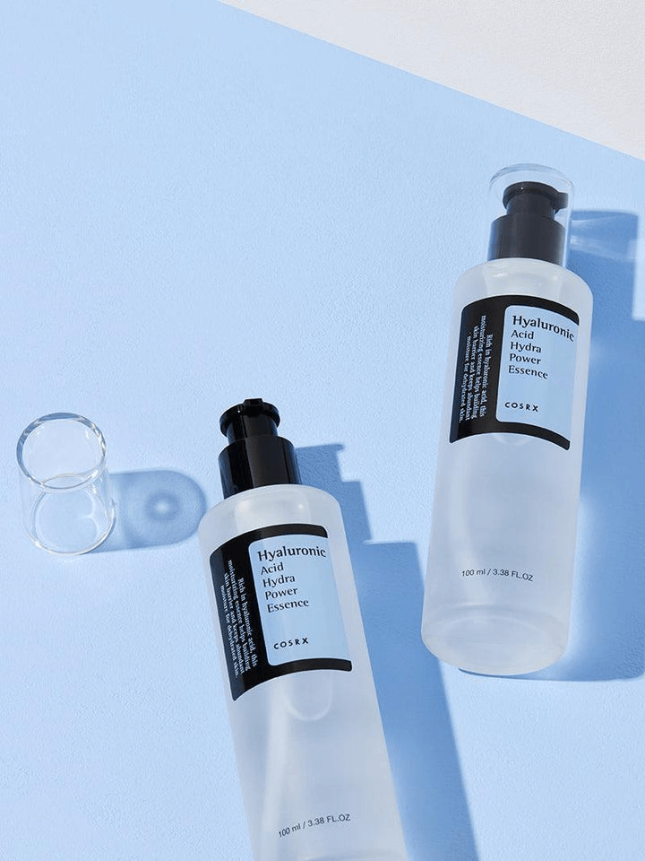 COSRX Hyaluronic Acid Hydra Power Essence bottles on a blue background, hydrating essence for dry skin.