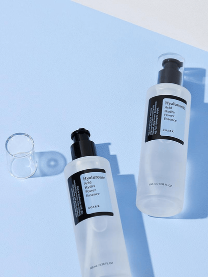 COSRX Hyaluronic Acid Hydra Power Essence bottles on a blue background, hydrating essence for dry skin.